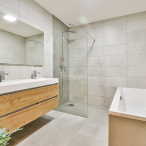 Badezimmer Impression modern, mit Doppelwaschbecken, ebenerdiger Regendusche und Badewanne, mit grauen Fliesen und Holzoptik
